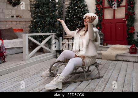Winterweihnachtsfrau, die auf einem Holzschlitten auf einer Veranda sitzt und die Winterweihnachtsszene genießt Stockfoto