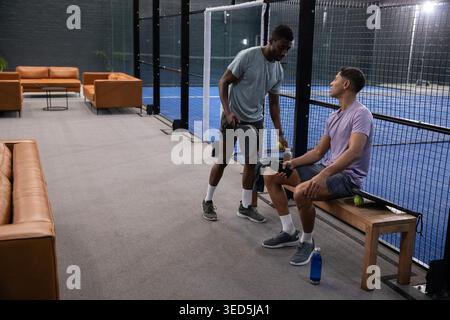 Verschiedene männliche Freunde in Sportbekleidung plaudern neben dem Padel-Platz auf der Bank, halten Paddel und Telefon Stockfoto