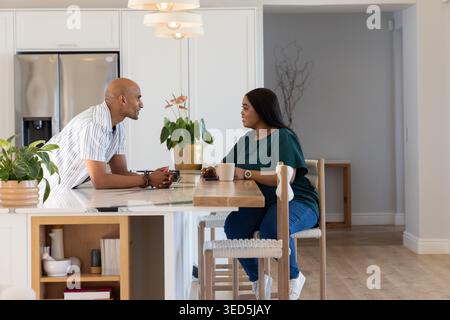 Ein Paar, das sich zu Hause über die Kücheninsel lehnt und sich bei Keramikbechern und Pflanzen unterhalten kann Stockfoto