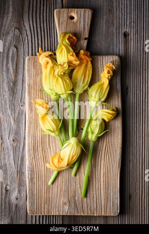 Zucchini-Blüten auf Schneidebrett mit rustikalem Hintergrund. Frische Speiseblumen mit Bio-Look und Kochzutaten-Konzept Stockfoto