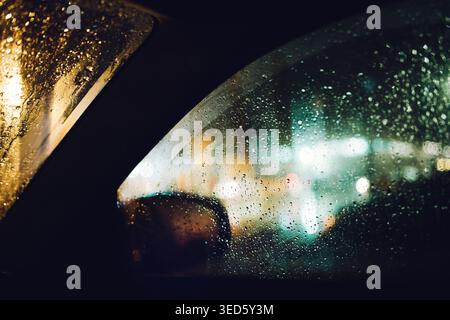 Regnerisches Nachtfenster mit City-Bokeh-Lichtern durch nasses Glas, selektiver Fokus Stockfoto