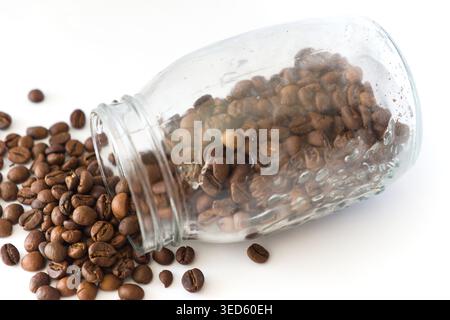 Ein transparenter Glasbehälter an der Seite, der geröstete Kaffeebohnen auf eine saubere weiße Oberfläche verschüttet, wodurch die reichhaltige Textur und Farbe der Bohnen hervorgehoben wird. Stockfoto
