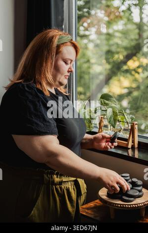 Frau im Profil, die Massagestein am Fenster mit Pflanze hält Stockfoto
