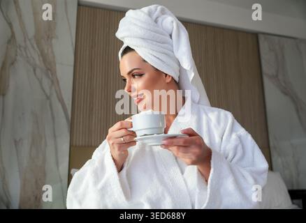 Eine Frau genießt eine Tasse Kaffee in einem Bademantel und trägt ein Handtuch auf dem Kopf. Frau steht in einem hellen Raum mit entspanntem Ausdruck. Die Stockfoto