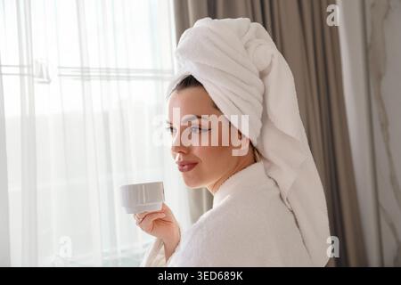 Eine Frau mit einem Handtuch auf dem Kopf hält eine weiße Tasse, während sie aus dem Fenster blickt. Die Frau trägt einen gemütlichen weißen Mantel. Helles Licht kommt von Th Stockfoto