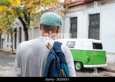 Rückansicht eines erwachsenen Mannes, der eine ruhige Wohnstraße mit Rucksack entlang geht und an einem Vintage-Van vorbeigeht. Stockfoto
