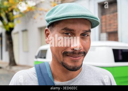 Mann mit grüner Mütze und T-Shirt, der selbstbewusst draußen auf einer Stadtstraße lächelt, mit einem Retro-Van im Hintergrund Stockfoto
