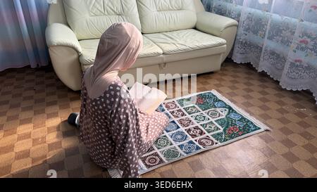 Junge Erwachsene muslimische Frau aus dem Nahen Osten in beigem Hijab und bescheidenem Kleid, die den Koran liest, während sie zu Hause auf einem Gebetsteg sitzt. Stockfoto