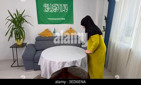 Junge Erwachsene muslimische Frau in schwarzem Hijab und gelbem Kleid, die zu Hause einen Tisch mit weißer Tischdecke für das Iftar-Essen während des Ramadan arrangiert. Stockfoto