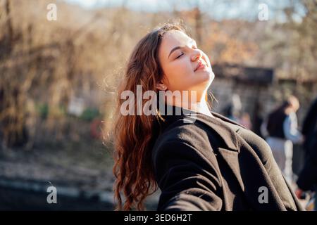 Junge kaukasische Frau mit langen Haaren in schwarzem Fell, die Sonnenlicht genießt und im Park die Augen geschlossen hat. Entspannung und Freizeit. Stockfoto