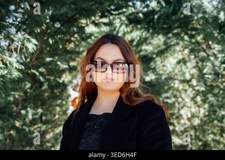 Junge kaukasische Frau mit langen braunen Haaren mit Sonnenbrille und schwarzer Jacke, die draußen im grünen Parkhintergrund steht. Modeporträt und Urban Stockfoto