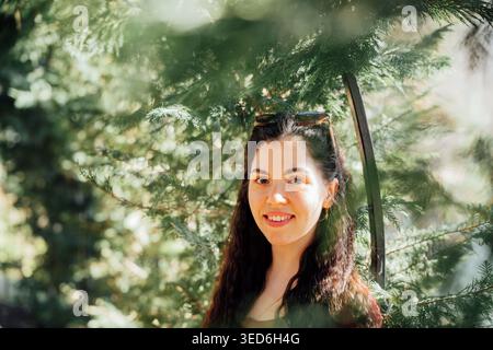 Junge kaukasische Frau mit langen dunklen Haaren lächelt und steht zwischen grünen Ästen im Park. Porträt im Freien und natürliches Lifestyle-Thema. Stockfoto