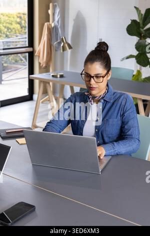 Frau mit Denim-Hemd und -Schal, die an einem silbernen Laptop am modernen Arbeitsplatz mit Smartphone arbeitet Stockfoto
