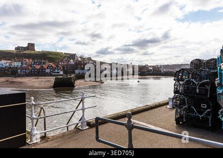 Blick auf Whitby, North Yorkshire, mit gestapelten Krabbentöpfen neben dem Hafen mit ruhigem Wasser, das in Richtung der Stadt führt. Reihen von Häusern mit rotem Dach erheben sich Stockfoto