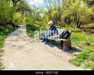 Älterer grauhaariger Mann, der auf einer Parkbank sitzt und sich darauf konzentriert, eine Drohne mit einem Handheld-Drohnencontroller zu bedienen. Sonniger Frühling. Pfad mit einem Mülleimer Stockfoto