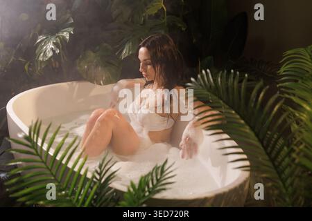 Eine ruhige Szene einer Frau, die ein Sprudelbad in einer stilvollen Badewanne genießt, umgeben von üppigem tropischem Laub, perfekt für Wellness- und Schönheitskampagnen. Stockfoto