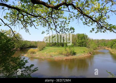 Ein ruhiger Fluss fließt durch eine lebendige grüne Landschaft, eingerahmt von überhängenden Ästen und einem hellen Himmel Stockfoto