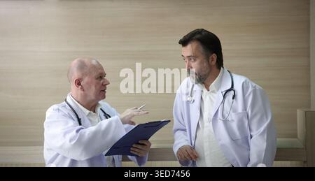 Ärzte diskutieren. Zwei erfahrene Ärzte diskutieren Patienteninformationen und medizinische Berichte in einem Krankenhauskorridor. Professionelle medizinische Teamarbeit c Stockfoto