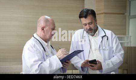Ärzte diskutieren. Zwei erfahrene Ärzte diskutieren Patienteninformationen und medizinische Berichte in einem Krankenhauskorridor. Professionelle medizinische Teamarbeit c Stockfoto