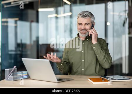 Ein reifer bärtiger Mann lächelt, telefoniert mit einem Laptop und Dokumenten am Schreibtisch, kommuniziert und verwaltet Aufgaben in einem modernen Arbeitsbereich Stockfoto