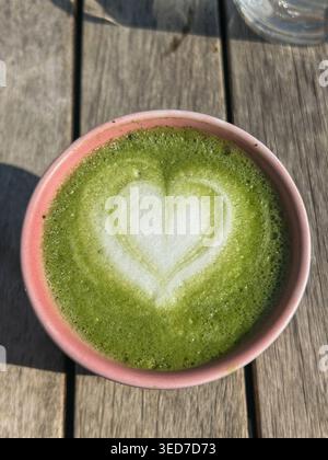 Blick von oben auf Matcha Latte mit herzförmiger Schaumkunst in einer Keramikschale auf einem Holztisch bei natürlichem Sonnenlicht. Stockfoto