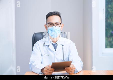 Porträt eines männlichen Arztes mit Gesichtsmaske und Labormantel, der am Schreibtisch sitzt Stockfoto