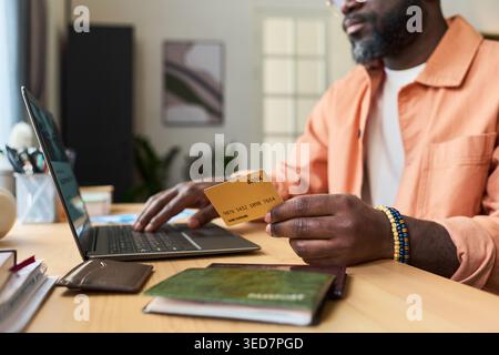 Schwarzer Mann mittleren Alters mit Bankkarte und Laptop bei der Vorbereitung der Reisebuchung mit Reisepass auf dem Schreibtisch, der Online-Zahlung und Reiseplanung für Reisemarketing darstellt Stockfoto