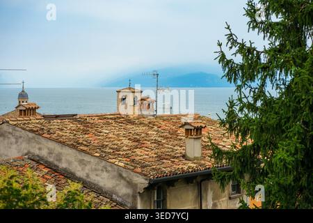 Alte, gekachelte Dächer mit Schornsteinen mit Blick auf den Gardasee in Italien, malerische Berglandschaft mit ruhigem Wasser und bewölktem Himmel, europäischer Reiseblick. Stockfoto