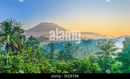 Nebliger tropischer Sonnenaufgang über üppigem Regenwald und Mount Agung Vulkan mit Palmen in Sideman, Bali Stockfoto