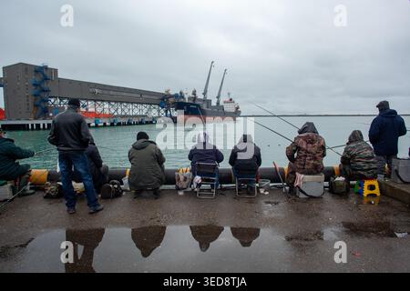 Tuapse Russland 21. Dezember 2015. Fischer sitzen am Hafenbarren-Fischen mit Reflexionen, die auf nassem Gehweg in der Nähe des Industriehafens sichtbar sind. Stockfoto