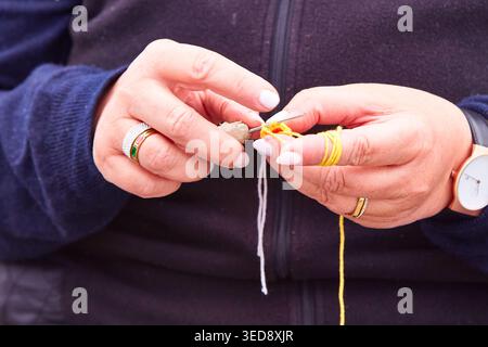 Frauenhände mit Ringen manipulieren eine Häkelnadel und ein gelbes Garn und zeigen Häkeltechnik, während sie einen dunklen Pullover aus nächster Nähe tragen Stockfoto