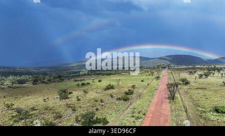 Aus der Vogelperspektive einer roten Feldstraße, die sich durch die grüne Savannenlandschaft unter einem doppelten Regenbogen und dunklem stürmischen Himmel erstreckt. Waterberg District Municipali Stockfoto