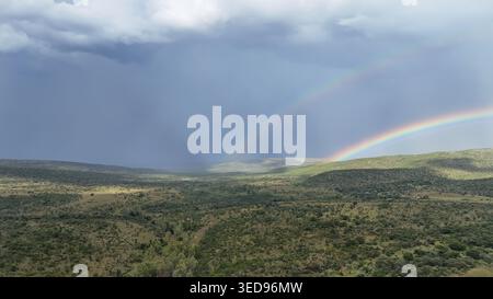 Aus der Vogelperspektive eines Doppelregenbogens, der sich über sanfte grüne Hügel und ein dichtes Buschland unter einem dramatischen stürmischen Himmel im Waterberg District Municipalit erstreckt Stockfoto