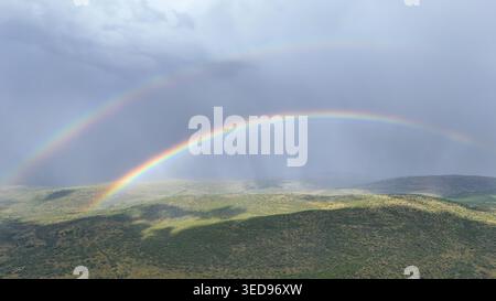 Aus der Vogelperspektive eines Doppelregenbogens über sanfte grüne Hügel und dichte Vegetation unter einem dramatischen bewölkten Himmel in Waterberg District Municipality, Limpopo, Stockfoto