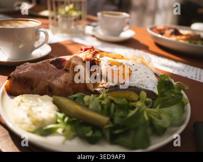Herzhafter Brunch-Teller mit Spiegeleiern, Speck, Salat und Gurke, serviert mit Kaffee im Café Ott and Matilda in Estland Stockfoto