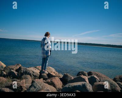 Junge Frau mit Sonnenbrille, die am felsigen Ufer am Lohusalu-Strand in Estland steht und die Frühlingssonne genießt Stockfoto