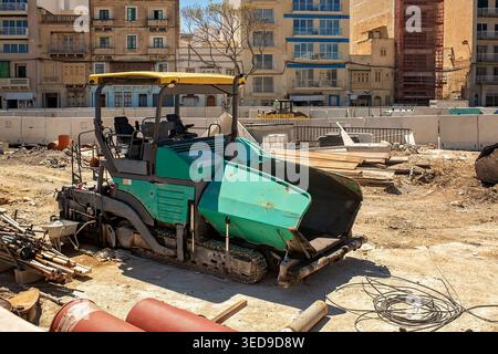 Auf einer stark frequentierten Straßenbaustelle in der Stadt steht ein grüner Asphaltfertiger. Ausbau der städtischen Infrastruktur und Straßenrenovierung in Malta. Stockfoto