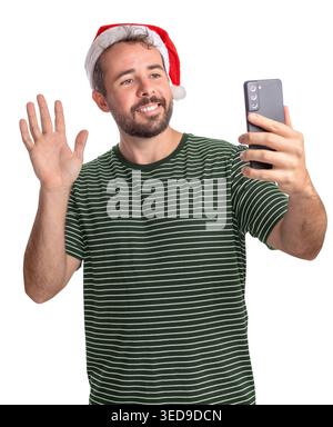 Ein bärtiger Mann trägt einen Weihnachtsmann-Hut. Er lächelt und winkt seinem Handy zu, während er einen Videoanruf macht. Die Umgebung ist hell und lässt ein festi vermuten Stockfoto