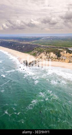 Der geschwungene Sandstrand von Praia da Paredes da Vitoria aus der Vogelperspektive. Türkisfarbenes Wasser prallt auf zerklüftete Felsen nahe der Küste. Weit entfernte Klippen erheben sich auf bes Stockfoto