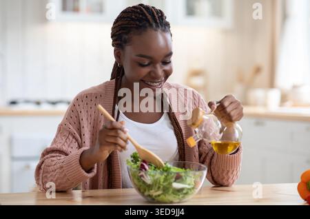 Lächelnde afroamerikanische Frau kocht Gemüsesalat in der Küche und fügt Olivenöl hinzu Stockfoto