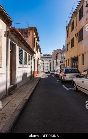 Enge europäische Straße auf Madeira, gesäumt von farbenfrohen Kolonialgebäuden und geparkten Fahrzeugen. Klarer blauer Himmel beleuchtet die Geschichte Stockfoto