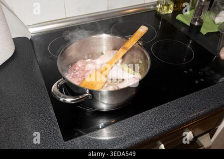 Geflügelstücke und Gemüse kochen in einem Stahltopf mit einem Holzlöffel auf einem Induktionskochfeld Stockfoto