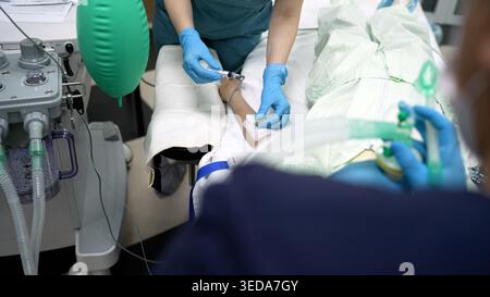 Die Krankenschwester in blauen Handschuhen injiziert Medikamente in die Infusionsleitung am Arm des Patienten. Medizinisches Team bereitet sich auf die Operation mit Anästhesiesystem im klinischen Krankenhaus vor Stockfoto