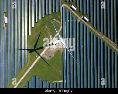 Luftaufnahme der Chelveston Wind Farm mit einer großen Windturbine, die einen Schatten über Reihen blauer Solarpaneele auf grünen Feldern in Wellingborough, Engla, wirft Stockfoto