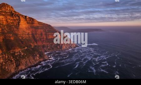 Aus der Vogelperspektive auf die zerklüfteten Klippen des Chapman's Peak Drive mit Blick auf den Atlantischen Ozean bei einem pulsierenden Sonnenuntergang mit Wellen, die gegen die Küste krachen Stockfoto