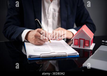Geschäftsmann unterschreibt Vertrag für den Kauf eines Hauses. Ein Mann, der unterschreibt, dass ein Hypothekarkreditvertrag mit dem Hausmodell des roten Dachs vorliegt. Immobilieninvestitionen, insu Stockfoto