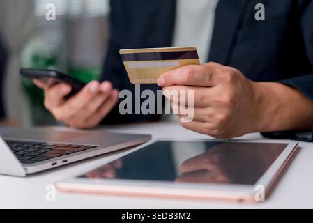 Nahaufnahme von Händen, die eine goldene Kreditkarte halten und ein Smartphone für Online-Shopping verwenden. E-Commerce-Konzept mit Laptop und Tablet auf dem Schreibtisch. Stockfoto