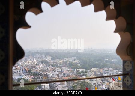 Eine Panoramblick auf eine dicht besiedelte indische Stadt, gesehen durch einen kunstvollen, muschelförmigen Bogenfensterrahmen. Im Vordergrund befindet sich ein dunkles Dekorelement Stockfoto