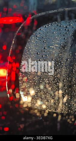 Nahaufnahme eines Seitenspiegels im Auto, bedeckt mit Regentropfen mit verschwommenen bunten Bokeh-Lichtern im Hintergrund. Stockfoto