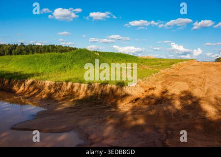 Ausgedehntes Sandgelände mit üppigem grünem Gras auf einem Hügel unter einem klaren blauen Himmel mit flauschigen Wolken, das eine ausgegrabene Fläche und einen unbefestigten Pfad zeigt Stockfoto